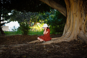 A cute european kid girl in dress and big hat sits near a big tree in the park. Child among large tree roots