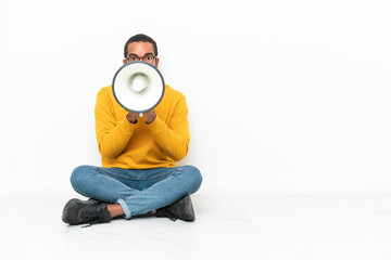 Young Ecuadorian man sitting on the floor isolated on white wall shouting through a megaphone