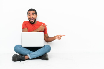 Young Ecuadorian man with a laptop sitting on the floor isolated on white background surprised and pointing side