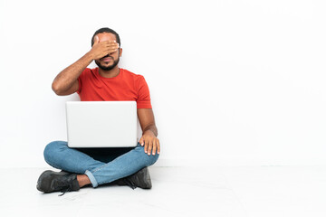 Young Ecuadorian man with a laptop sitting on the floor isolated on white background covering eyes by hands. Do not want to see something