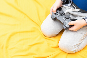 Black joystick for playing the video game in child hands on yellow fabric background. Playing video games at home concept. Spending time together. Top view with copy space