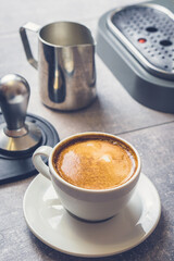 White cup with fresh hot coffee and milk on table near espresso machine pitcher and tamper