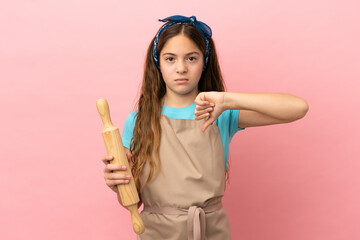 Little caucasian girl holding a rolling pin isolated on pink background showing thumb down with negative expression