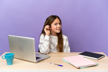 Little student girl in a workplace with a laptop isolated on purple background listening to something by putting hand on the ear