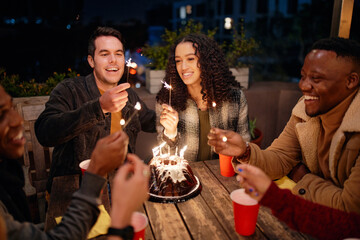 Group of diverse young adults sitting at boho themed rooftop party lighting sparklers from birthday cake.