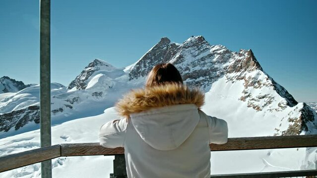 Tourist Girl looking at The summit of Jungfrau from the Sphinx Observatory. A young woman in a winter puffer jacket enjoying a Panoramic view of the highest summits of the Swiss Alps on a sunny day.