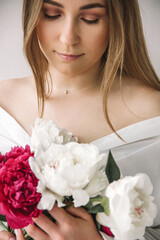 closeup young blonde woman in white shirt is standing with peonies in hands and looking down near white wall background, free space