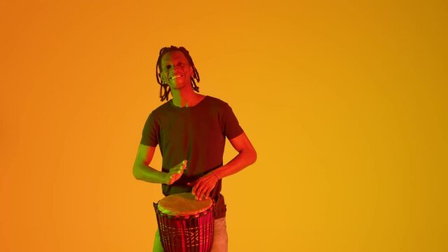 African American funky man in modern clothes cheerfully and energetically plays traditional African drum in background orange neon lit studio. Black man plays the bongo