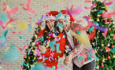 Young Caucasian lover couple husband and wife in red Santa Claus hat and sweater standing laughing holding shooting paper cracker confetti together in decorated living room with Christmas eve tree