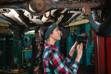 Fototapeta na wymiar Portrait of a young female mechanic in uniform who uses a tablet to diagnose a car. The car is on the lift. Side view
