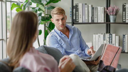 Millennial lover couple handsome husband in casual outfit sitting on sofa watching streaming online movie via notebook while beautiful wife using laptop computer working typing report during pandemic