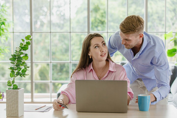 Millennial happy Caucasian female professional businesswoman entrepreneur working with laptop computer at home office and husband stand beside in living room.