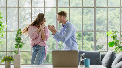 Millennial young Caucasian lover couple handsome husband and beautiful wife in casual outfit listening to music song playlist from laptop notebook computer dancing together in living room at home