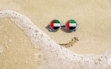 A painted smile on the sand and sunglasses with the flag of the UAE. The concept of a positive and successful holiday in the resort of the United Arab Emirates.