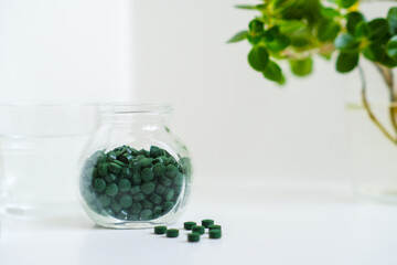 Green round spirulina pills in a jar with glass of water on a simple minimalistic white table. Bright houseplant in a frame. Superfood. Seaweed health care. Vitamin nutrition and treatment.