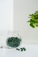 Green round spirulina pills in a jar with glass of water on a simple minimalistic white table. Bright houseplant in a frame. Superfood. Seaweed health care. Vitamin nutrition and treatment.