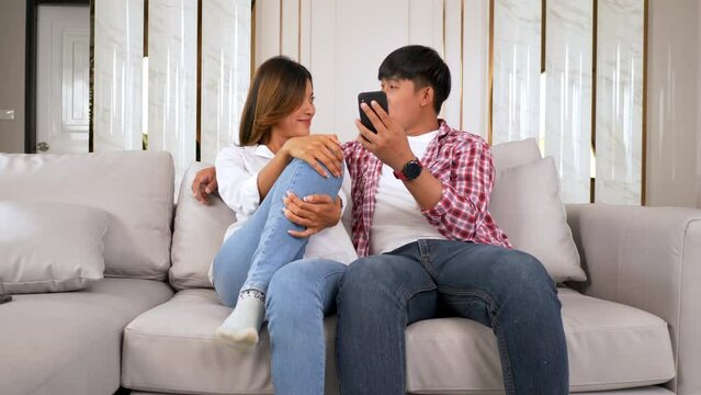 Slow motion shot, Happy Young couple sitting on sofa in living room at home use smartphone chatting with friend, talking and laughing together with happiness, happy family concept
