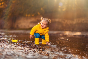 A funny smiling little girl in yellow rubber boots is playing with a paper boat by the river. Happy childhood
