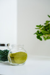 Green round spirulina pills and herbal detox powder in a jar on simple minimalistic white table, houseplant in a frame. Superfood. Seaweed health care. Vitamin nutrition and treatment. Immune system.