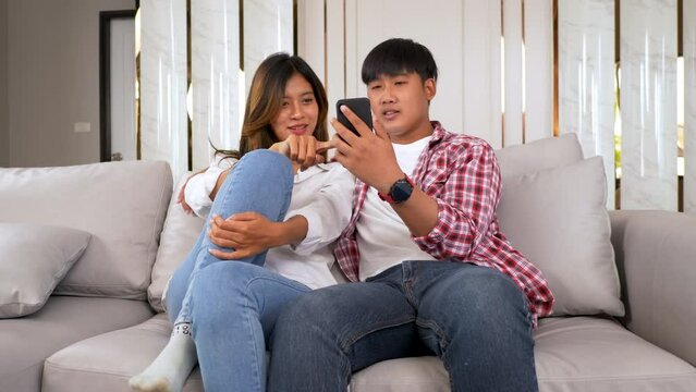 Slow motion shot, Young couple sitting on sofa in living room and use smartphone playing game together, they talk and laughing with happiness, happy family concept