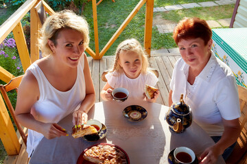Little blonde girl, mother and grandmother at a table on wooden terrace on nature or in the village and a green landscape in the background on a summer or autumn sunny day. Concept happy family