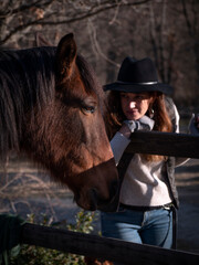 Trendy female with long hair, black hat and wool mittens looking at a chestnut mare.