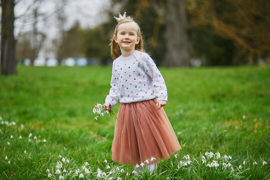 Cute preschooler girl in princess crown standing in the grass with many snowdrop flowers in park or forest on a spring day