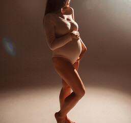 pregnant body. pregnant woman with red hair is standing in warm light in studio with hands on...