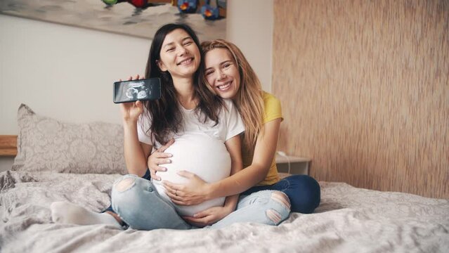 Beautiful pregnant woman holding and looking ultrasound photo newborn baby together with girlfriend on bed in bedroom. Family, woman love and romantic relationship. Beautiful asian friend in home