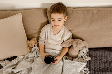 Adorable little boy drinking hot tea on the bed at home and relaxing. Child resting, wrapped in a blanket, with mug in his room. Interior and clothes in natural earth colors. Cozy environment.