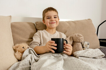 Adorable little boy drinking hot tea on the bed at home and relaxing. Child resting, wrapped in a blanket, with mug in his room. Interior and clothes in natural earth colors. Cozy environment.