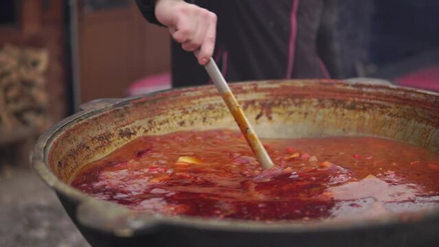 Cooking hungarian soup bograch on fire in a big cauldron outside with man stirring it with a ladle