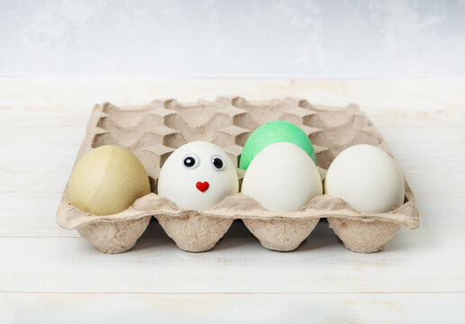 The egg carton contains dyed and white eggs. One egg has a cute face. Easter concept