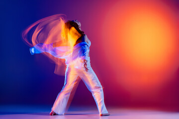 Image of flexible young girl, hip-hop dancer in white outfit dancing hip hop isolated on blue background in yellow neon light. Youth culture, style and fashion, action.