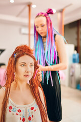 Female hairdresser with colored afro braids weaves to redhead girl ginger dreadlocks. Beauty salon services. Hippie and boho style coiffure with kanekalon.