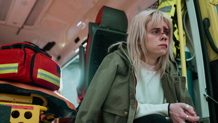 Caucasian Female Sitting at the Back of an Ambulance Car Waiting For Help From Medics. Face Covered in Bruises and Blood As She Looks Sad, War Survivor With Wounds All Over The Face.