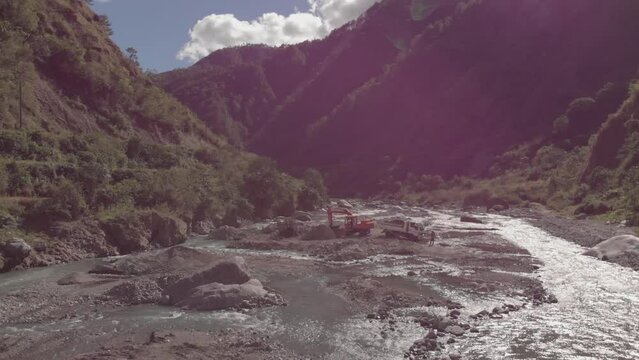 excavator digging rocks out of river bed in mountainous region in Benguet Kabayan Philippines wide angle aerial approaching raw cement material mining flowing water reflecting light bright sunny day