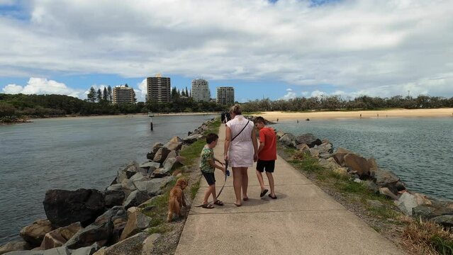 Two playful young brothers play tag around their mum who walks a dog along a rock wall pathway towards the Spit beach, Sunshine Coast.