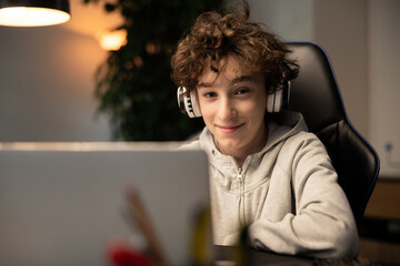 A young boy with curly hair is happy with remote learning. A student sits in front of a computer with wireless headphones listening to a teacher lecture. The lessons take place over the Internet.