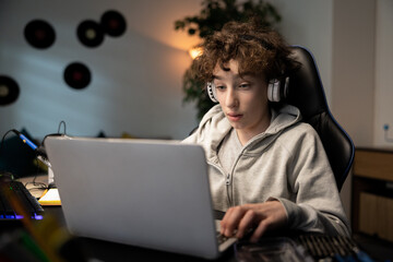 Boy at young age sits at desk in room in front of laptop, watches videos, listens to music through headphones, spends free time on internet.