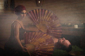 A woman performs a healing ritual with a Chinese fan for a man. A man lies on spruce branches