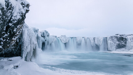 Goðafoss waterfall in winter. Ice formations around falls and snow on ground. Overcast sky. North Iceland.