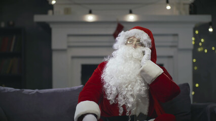 Santa Claus talking on smartphone sitting on couch in decorated living room