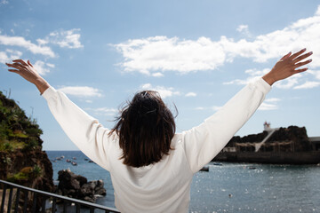 behind carefree young woman with arms raised by seaside