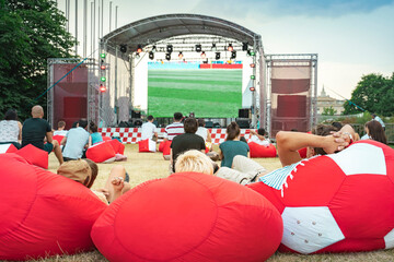 Outdoor event group of people watching soccer game on outdoor screen in city park on summer evening