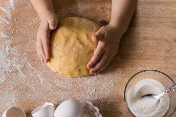Top view of human children's hands kneading dough on a wooden table in the kitchen. A child helping his mother to cook a pie kneading the dough in the kitchen.