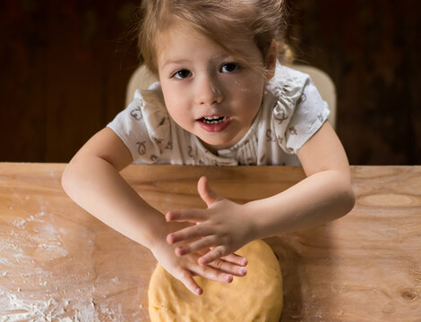 Top View Of Happy Blondy Girl Looking At Camera, Kneading Dough With Flour And Eggs On A Wooden Table, Helping Her Mother To Cook A Pie Kneading The Dough  In The Light Kitchen