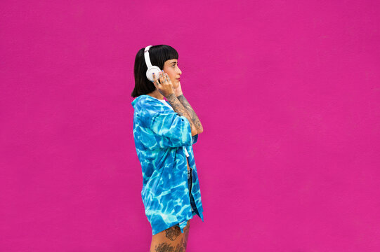 Joven mujer caucásica con cuerpo tatuado escuchando música con un auriculares sobre un fondo rosa