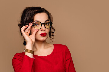 Pretty young woman wear red dress and round stylish glasses posing in studio over beige background....