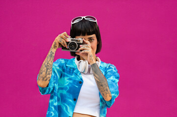 Joven mujer caucásica con cuerpo tatuado usando una cámara vintage sobre un fondo rosa